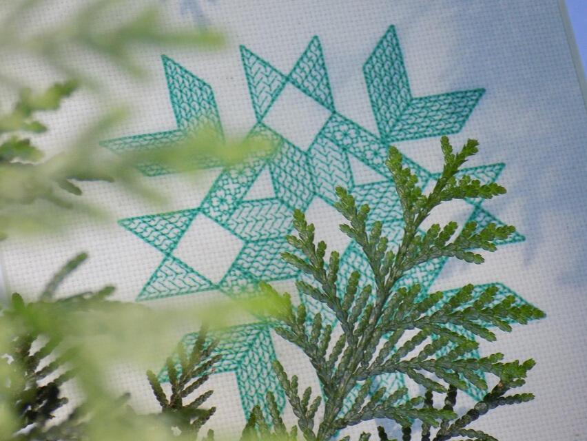 a piece of modern blackwork embroidery done in green thread sitting behind some ferns